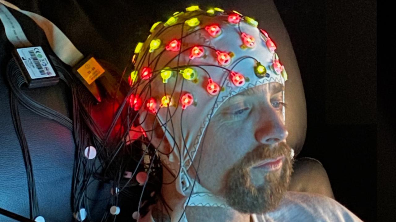 A man sits in a chair, wearing a lit up EEG cap. 